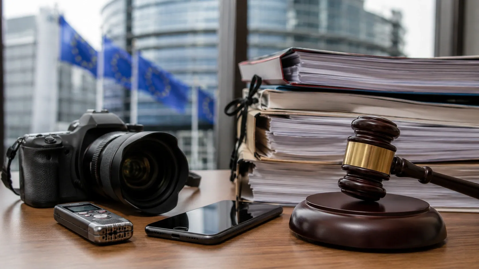 Camera, smartphone, recorder, judge's gavel, and binders with documents lay on the table with the EU flags in the background behind the window