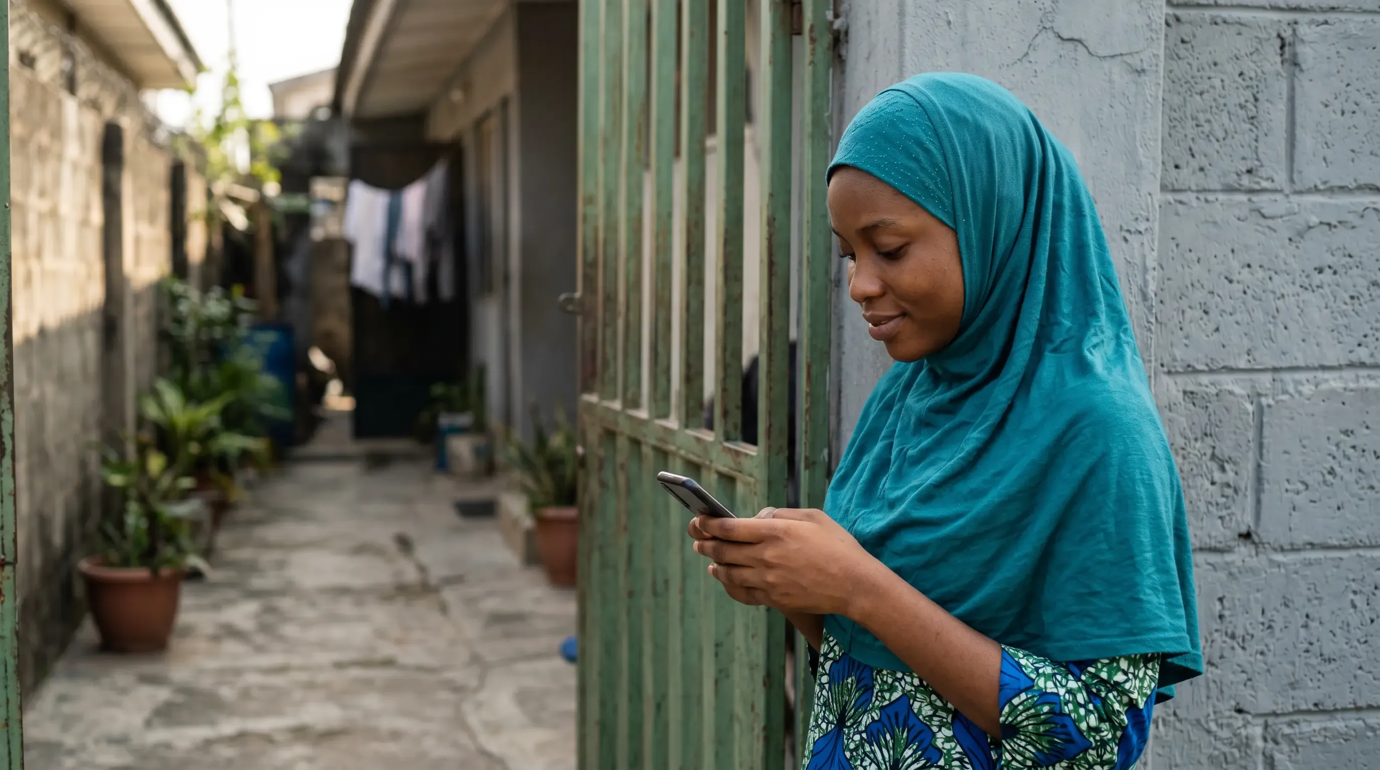 An image of a Nigerian woman in a hijab using her phone outside of her house